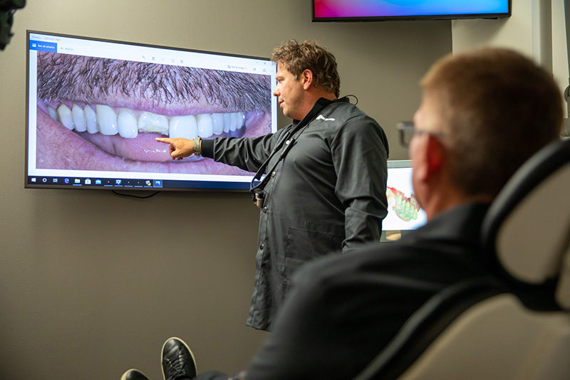 Prosthodontist Dr. Dean Kois pointing at a monitor showing a broken tooth and male patient siting in a dental chair looking on.