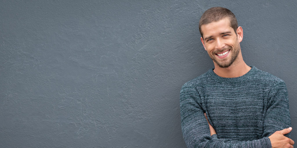 Man with 5 o'clock shadow and short hair standing in front of solid gray wall.