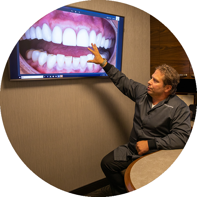 Prosthodontist Dr. Dean Kois sitting and pointing at a monitor showing a mouth with beautiful porcelain veneeers.
