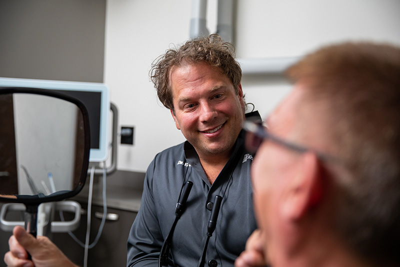 Prosthodontist Dr. Dean Kois showing male patient results of porcelain veneer treatment in a mirror.