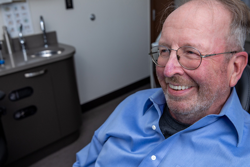 Elderly male in glasses and blue shirt smiling, showing his full mouth dental implants.